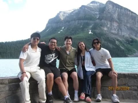 Five friends sitting on a stone wall in front of a turquoise lake and mountains.
