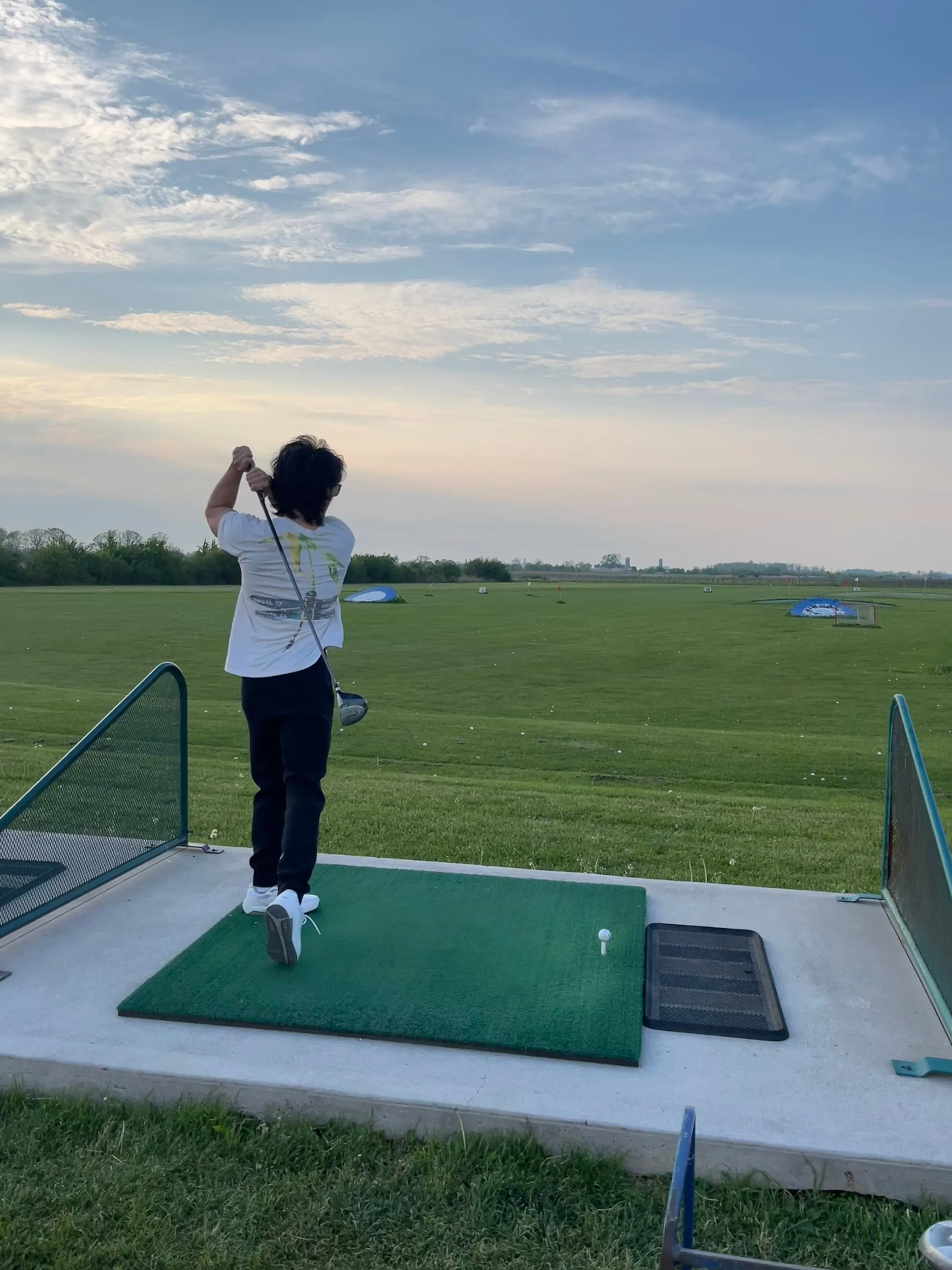 A golfer mid-swing at a driving range under an evening sky.